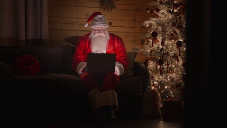 Der-Weihnachtsmann-Arbeitet-Am-Heiligabend-Zu-Hause-Mit-Einem-Laptop.-Weihnachtsbeleuchtung-Und-Dekoration